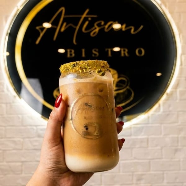 Hand holding an iced coffee with pistachio rim, in front of a glowing "Artisan Bistro" sign.