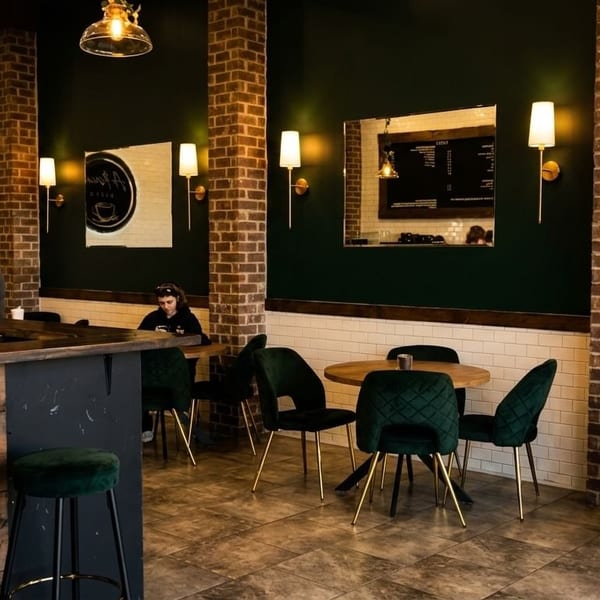 A cozy coffee shop interior with a barista, dark green walls, brick pillars, and velvet seating.