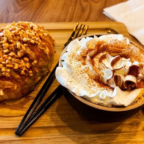 A croissant topped with nuts and a coffee with whipped cream and cinnamon on a wooden tray.