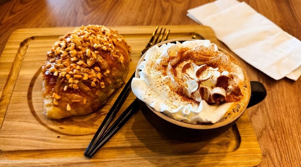 A croissant topped with nuts and a coffee with whipped cream and cinnamon on a wooden tray.