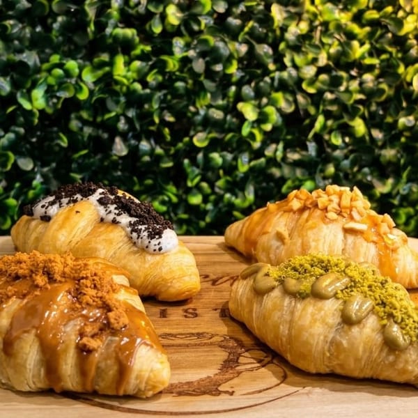 Four gourmet croissants with various sweet toppings on a wooden board against a green leafy background.