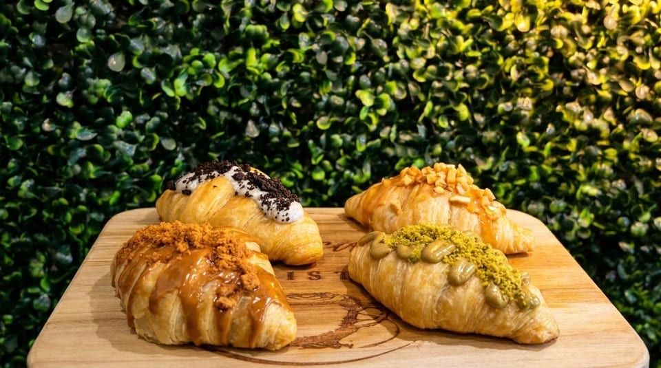 Four gourmet croissants with various sweet toppings on a wooden board against a green leafy background.