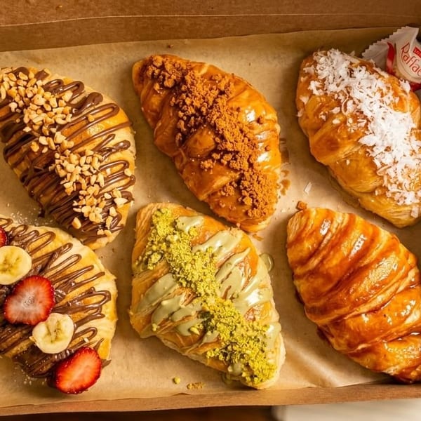 Six gourmet croissants with various toppings like nuts, fruit, and coconut, in a brown box.